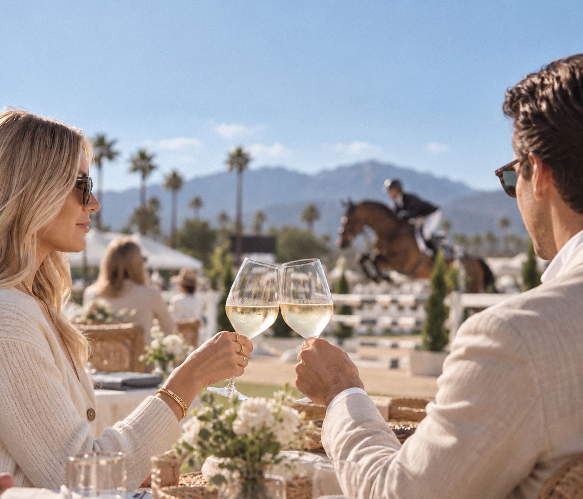Two guests toasting with white wine as a rider clears a fence beneath the San Gabriel Mountains